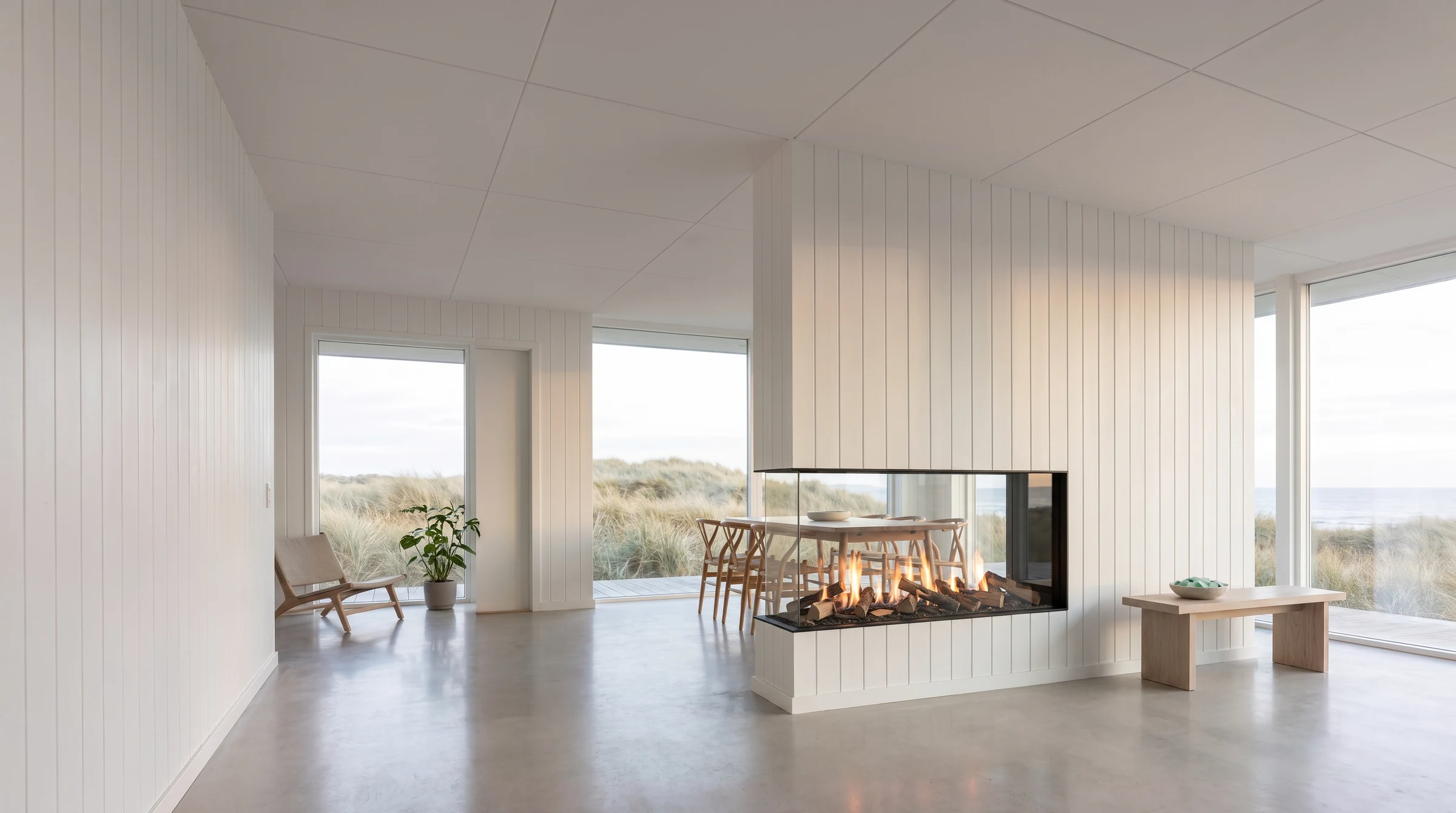 Chimenea de gas Fuegolo FGH1 U-line 1400 en una pared blanca de paneles de una casa junto al mar, con llamas entre troncos cerámicos y dunas al otro lado de las ventanas
